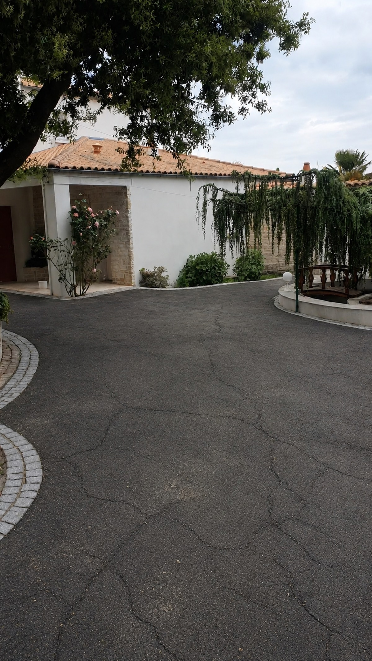 Cour en bitume craquelé et fissuré devant une maison de caractère, mauvaises herbes dans les joints